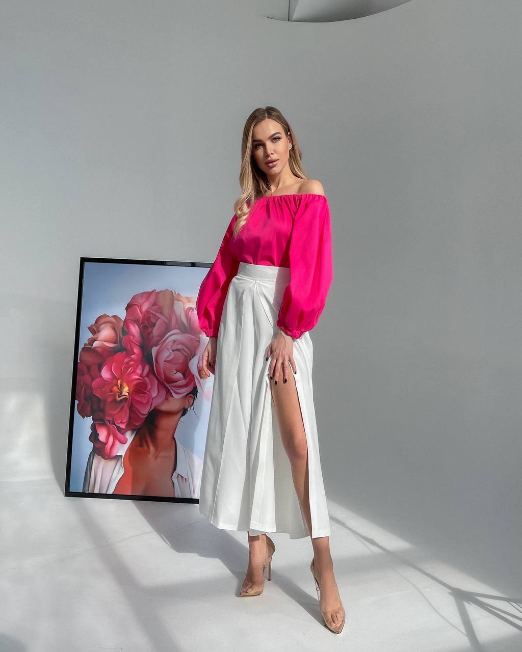 Woman in a bright pink top and white skirt standing in front of a floral painting.