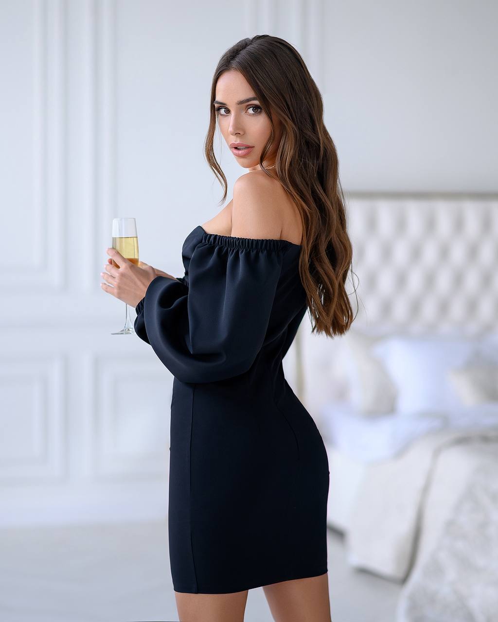 Woman in a black off-shoulder dress holding a glass of champagne in a room with white walls.