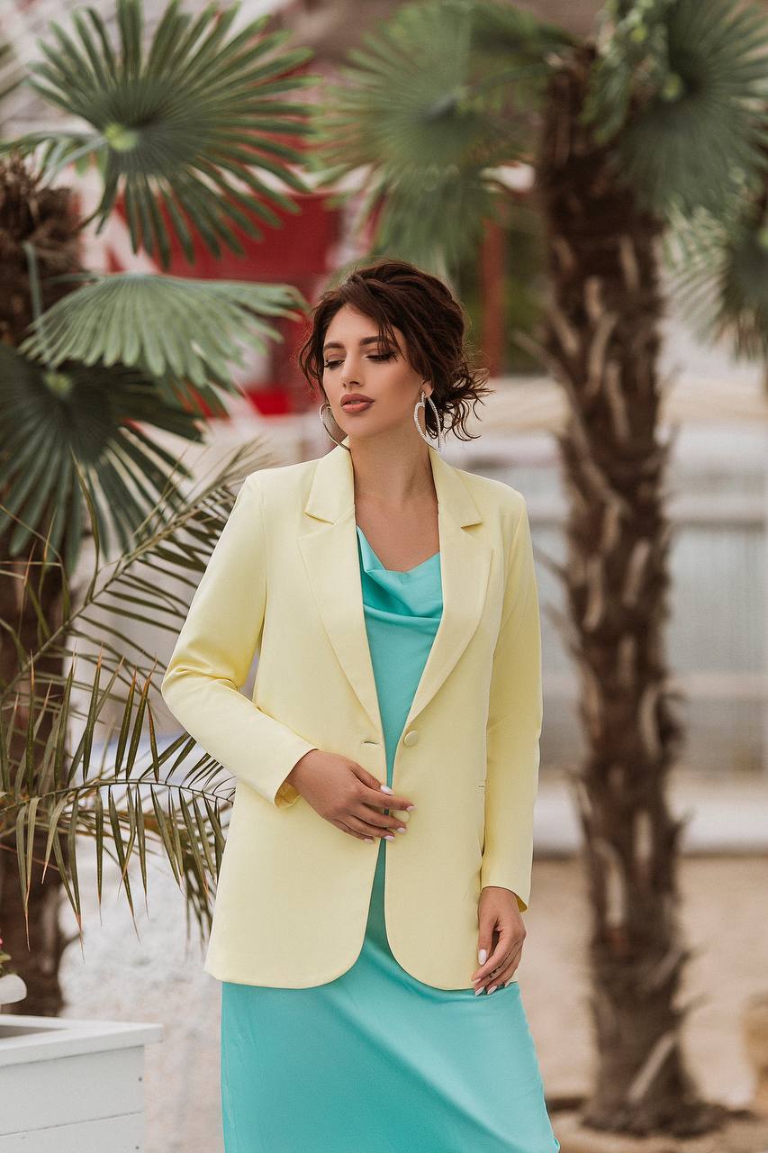 A woman wearing a pale yellow blazer over a turquoise dress, standing outdoors in front of palm trees.