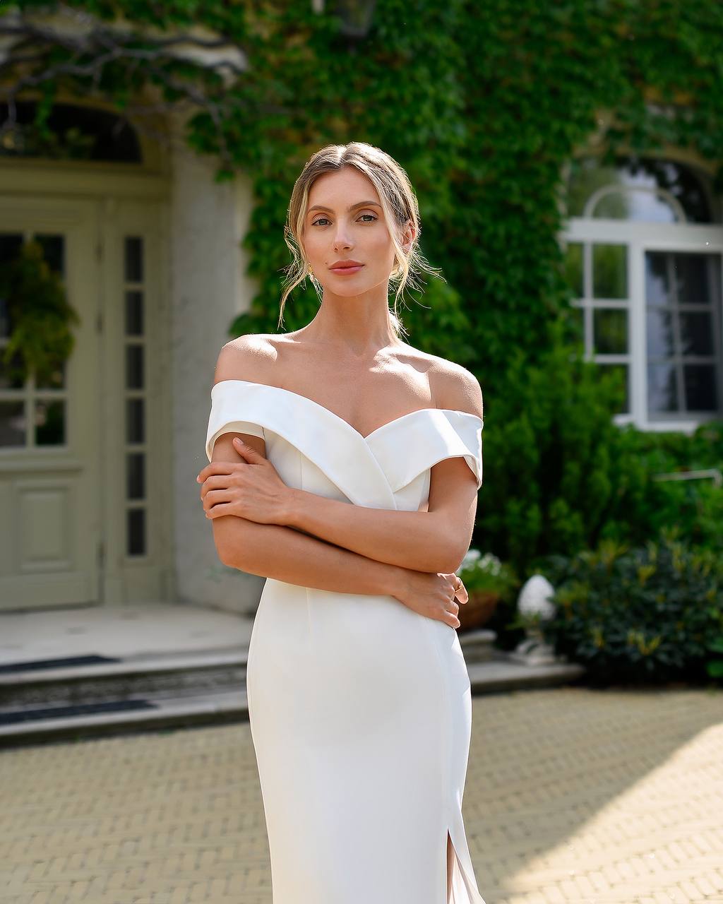 Woman in a white off-shoulder dress standing outdoors with greenery and a building in the background