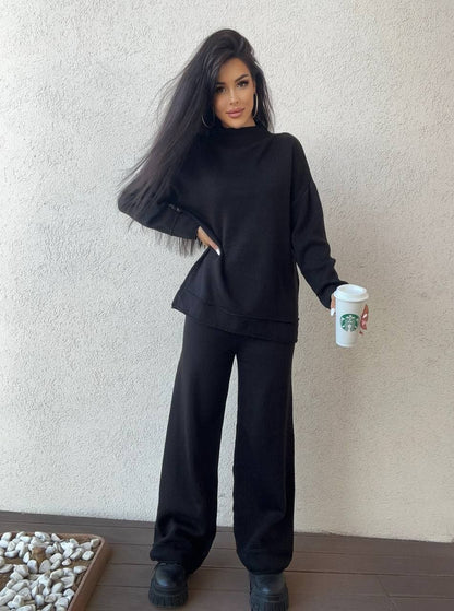 Person wearing a black outfit holding a Starbucks cup against a plain wall.