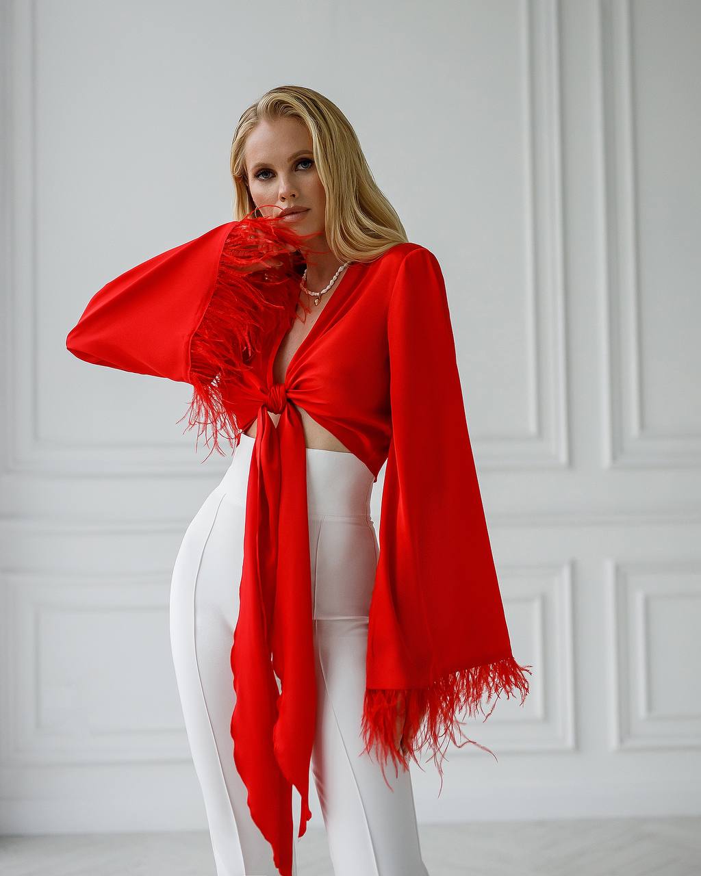 Woman wearing a red scarf and white pants against a light gray wall.