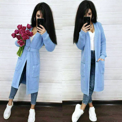 Woman wearing a long blue cardigan holding pink flowers in front of a white wall.