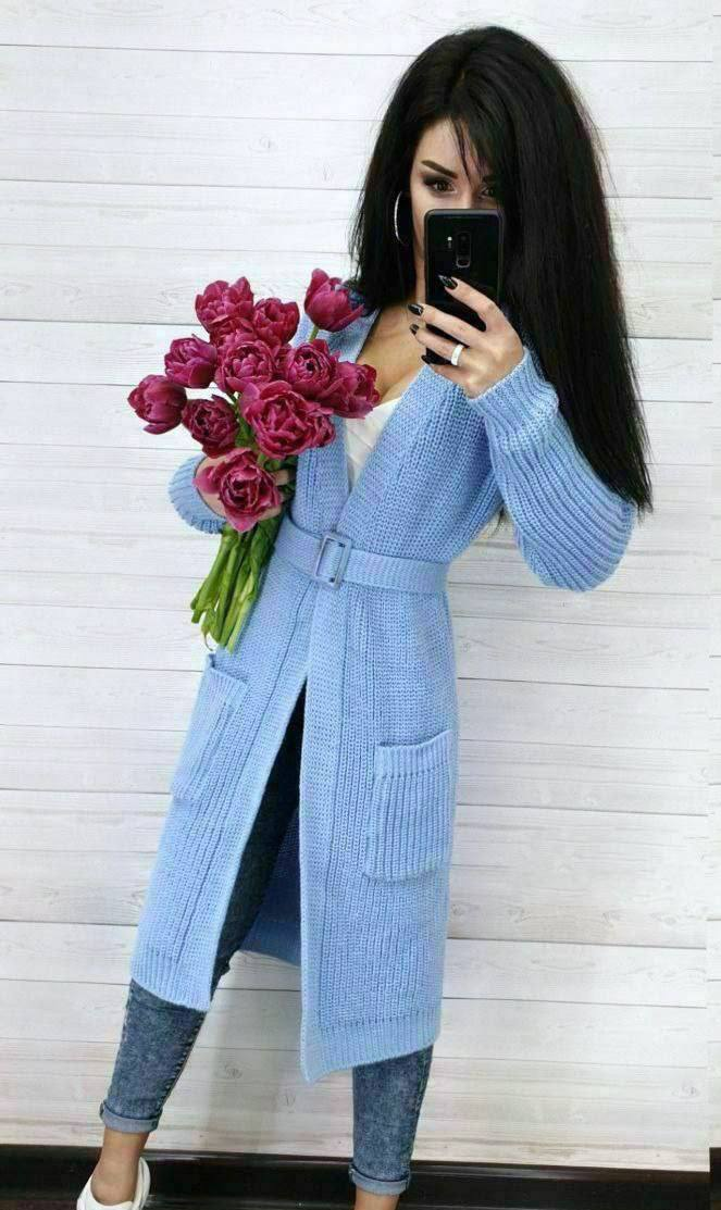 Woman wearing a long light blue cardigan holding pink roses against a white wall.