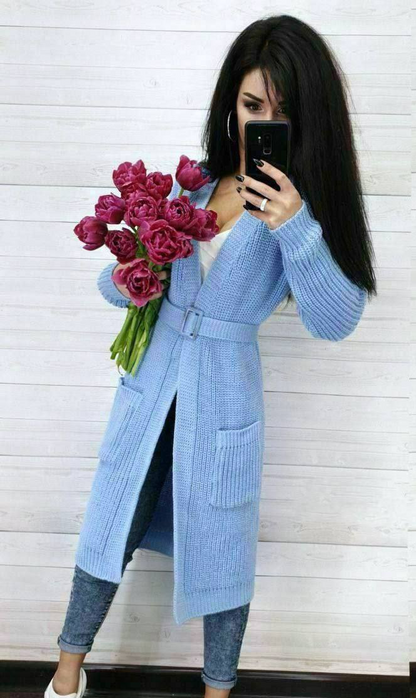 Woman wearing a long light blue cardigan holding pink roses against a white wall.