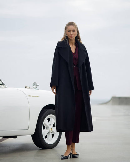Woman in a long black coat standing next to a white car in a minimalistic setting
