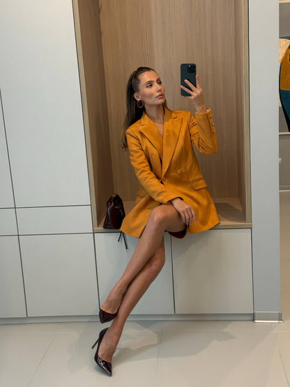 Woman in a mustard yellow coat taking a mirror selfie in a store.