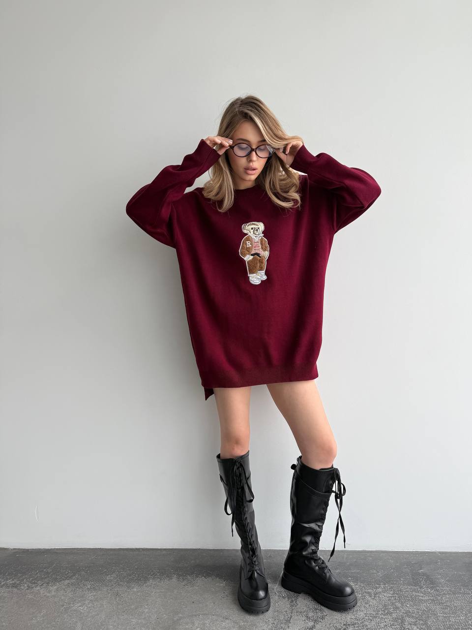 Person wearing a burgundy oversized sweater with a graphic design and black boots against a plain background