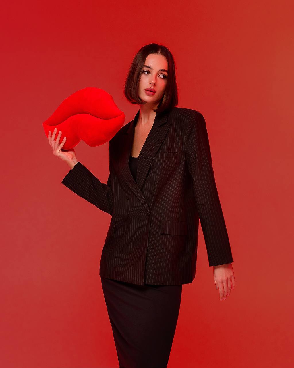 Woman in a black suit holding a large red lips prop against a red background