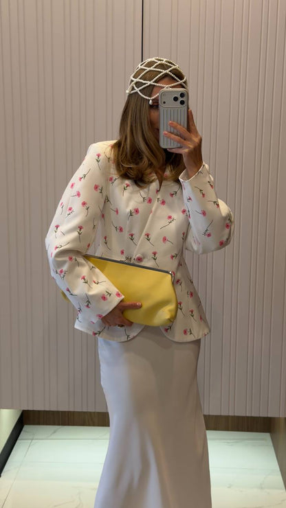 Person wearing a white floral blouse and holding a yellow clutch in front of a striped wall.