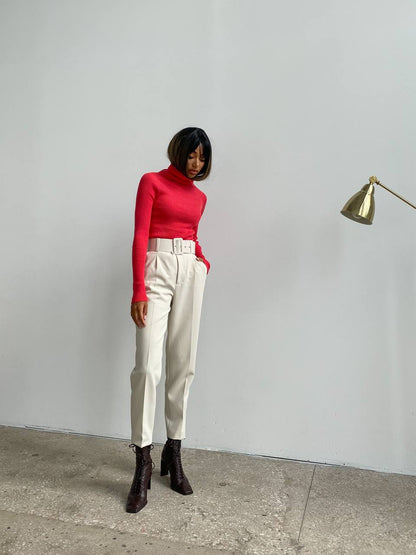 Person wearing a red turtleneck and white pants standing against a plain wall.