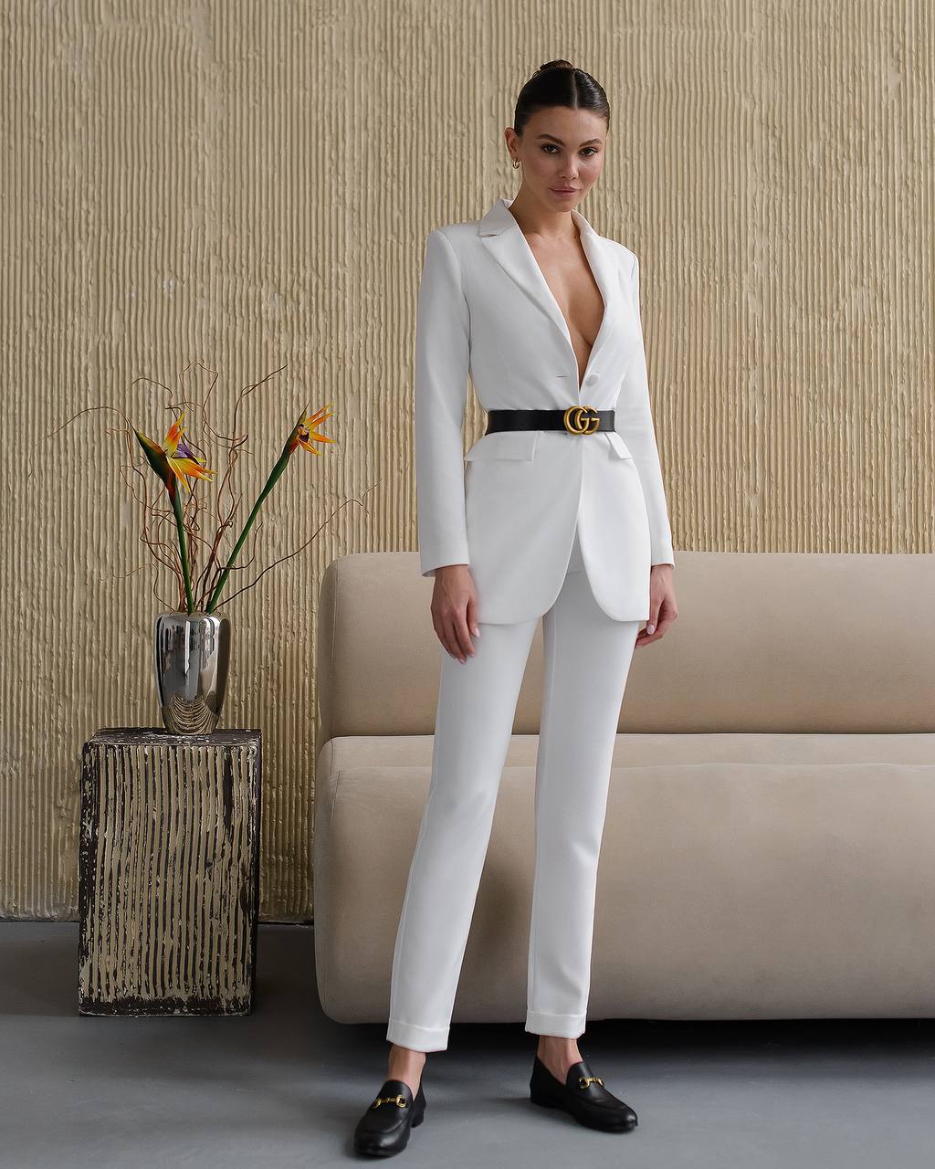 Woman in a white suit standing in a room with a beige sofa and decorative elements.