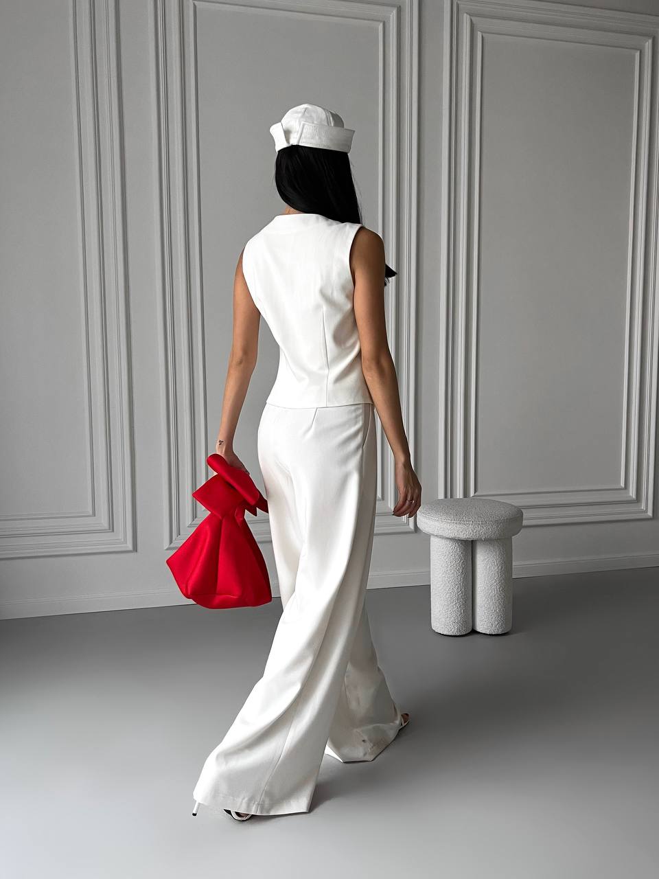 Person wearing a white outfit with a red bag in a minimalistic room.
