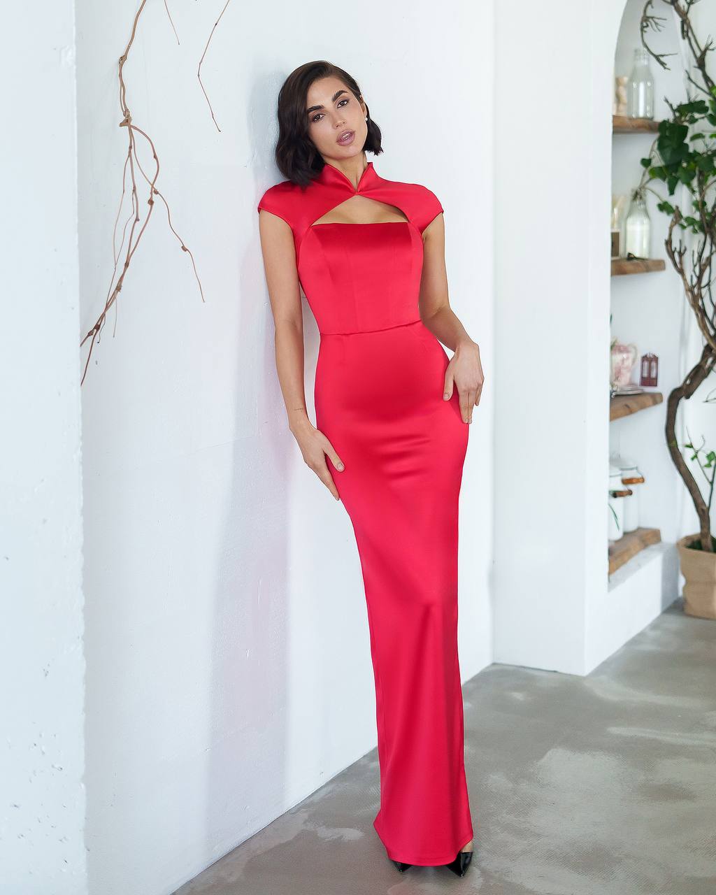 Woman wearing a red dress standing against a white wall with a plant in the background.