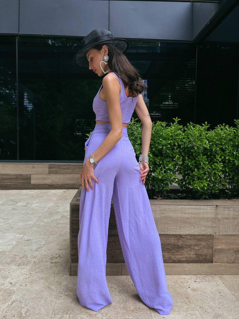 Woman in a lavender outfit standing outdoors with a dark wall and greenery in the background