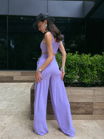 Woman in a lavender outfit standing outdoors with a dark wall and greenery in the background