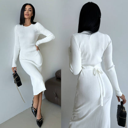 Woman wearing a white ribbed dress with long sleeves and a belt, holding a black handbag.