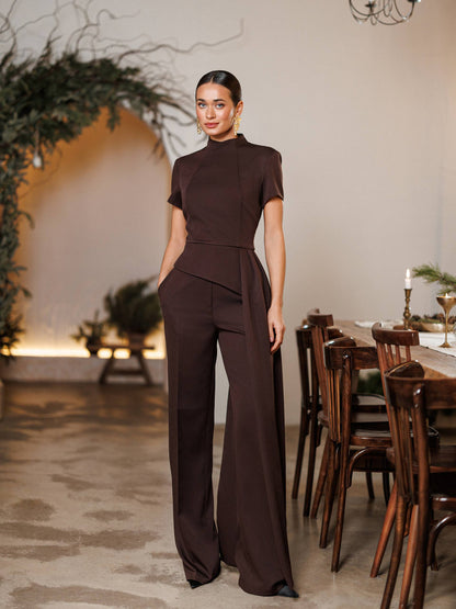 Woman in a brown jumpsuit standing in a stylishly decorated room with wooden furniture and plants.