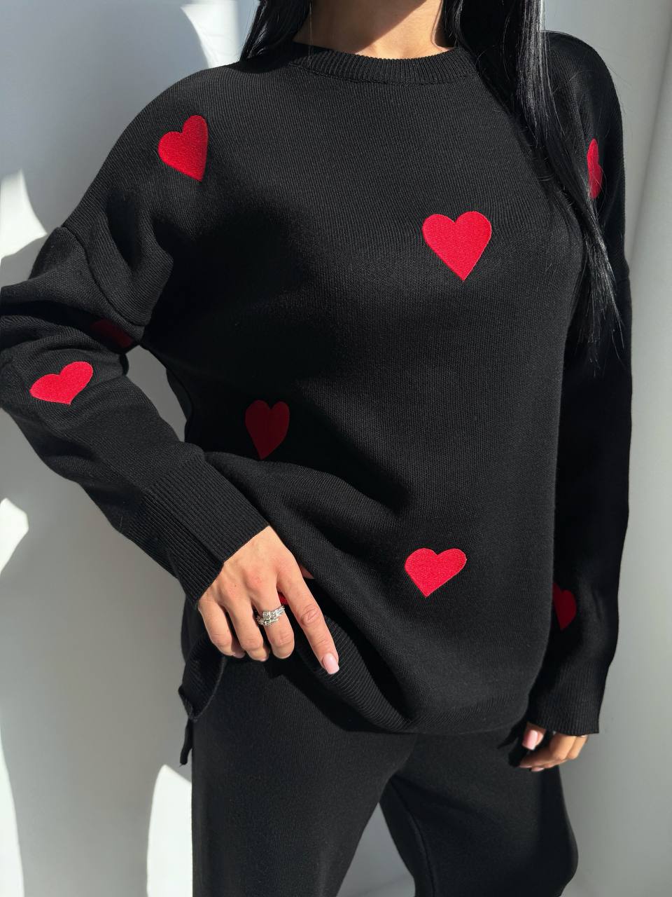 Black sweater with red heart patterns worn by a person against a neutral background