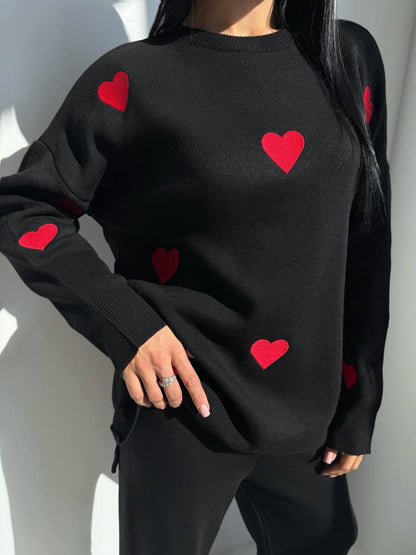 Black sweater with red heart patterns worn by a person against a neutral background