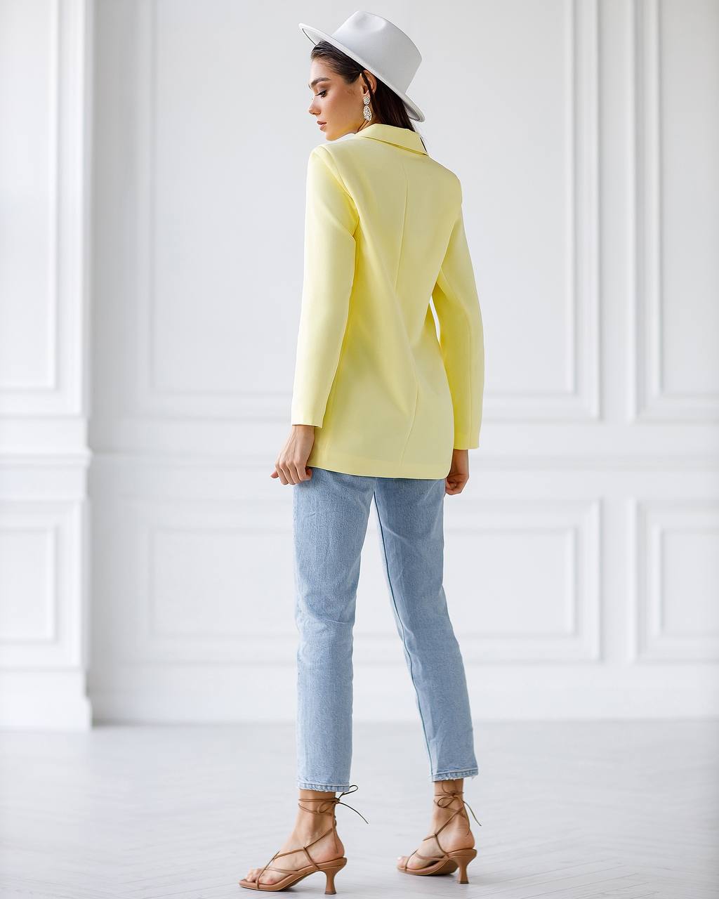A woman shown from the back wearing a light yellow blazer, light blue jeans, and tan lace-up heeled sandals in a bright white room with decorative wall paneling.