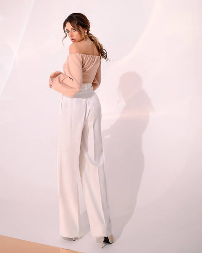 Woman wearing a beige off-shoulder top and white pants against a white background