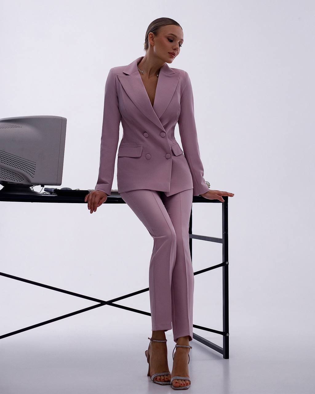 Woman in a pink suit standing next to a desk with a computer monitor on a white background