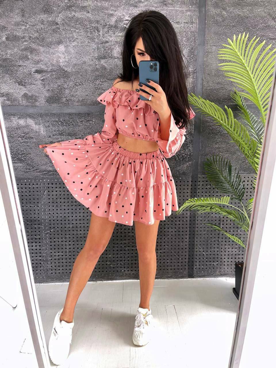 Woman taking a mirror selfie wearing a pink polka dot outfit with a gray wall and plant in the background.