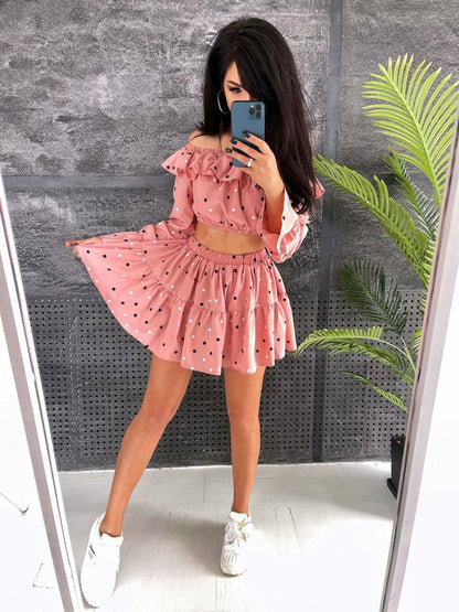 Woman taking a mirror selfie wearing a pink polka dot outfit with a gray wall and plant in the background.