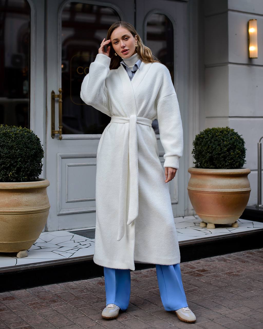 Woman in a white robe standing outside a building with potted plants.