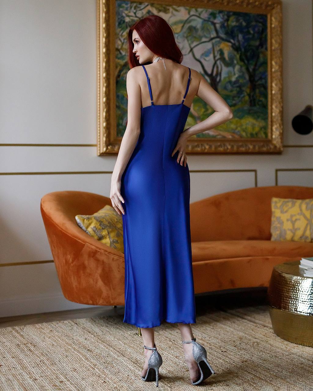 Woman in a blue dress standing in a stylish room with an orange sofa and gold accents.