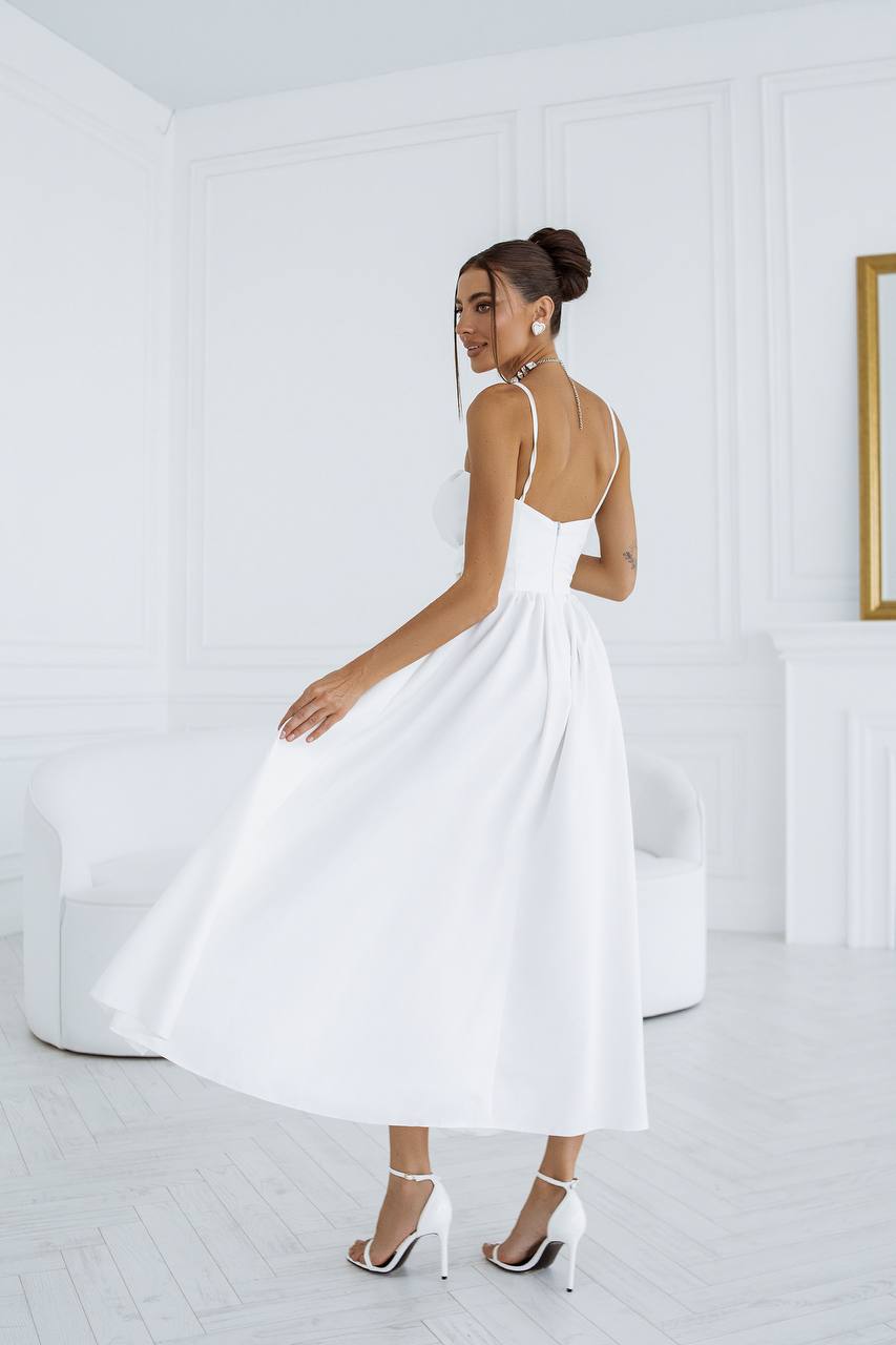 Woman in a white dress standing in a minimalistic room with a white wall and light-colored floor.