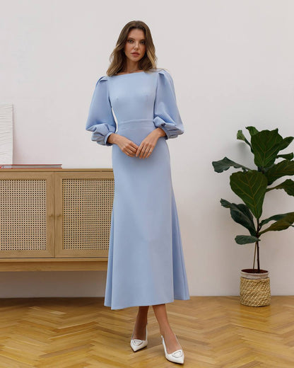 Woman wearing a light blue dress standing in a room with a wooden cabinet and plant.