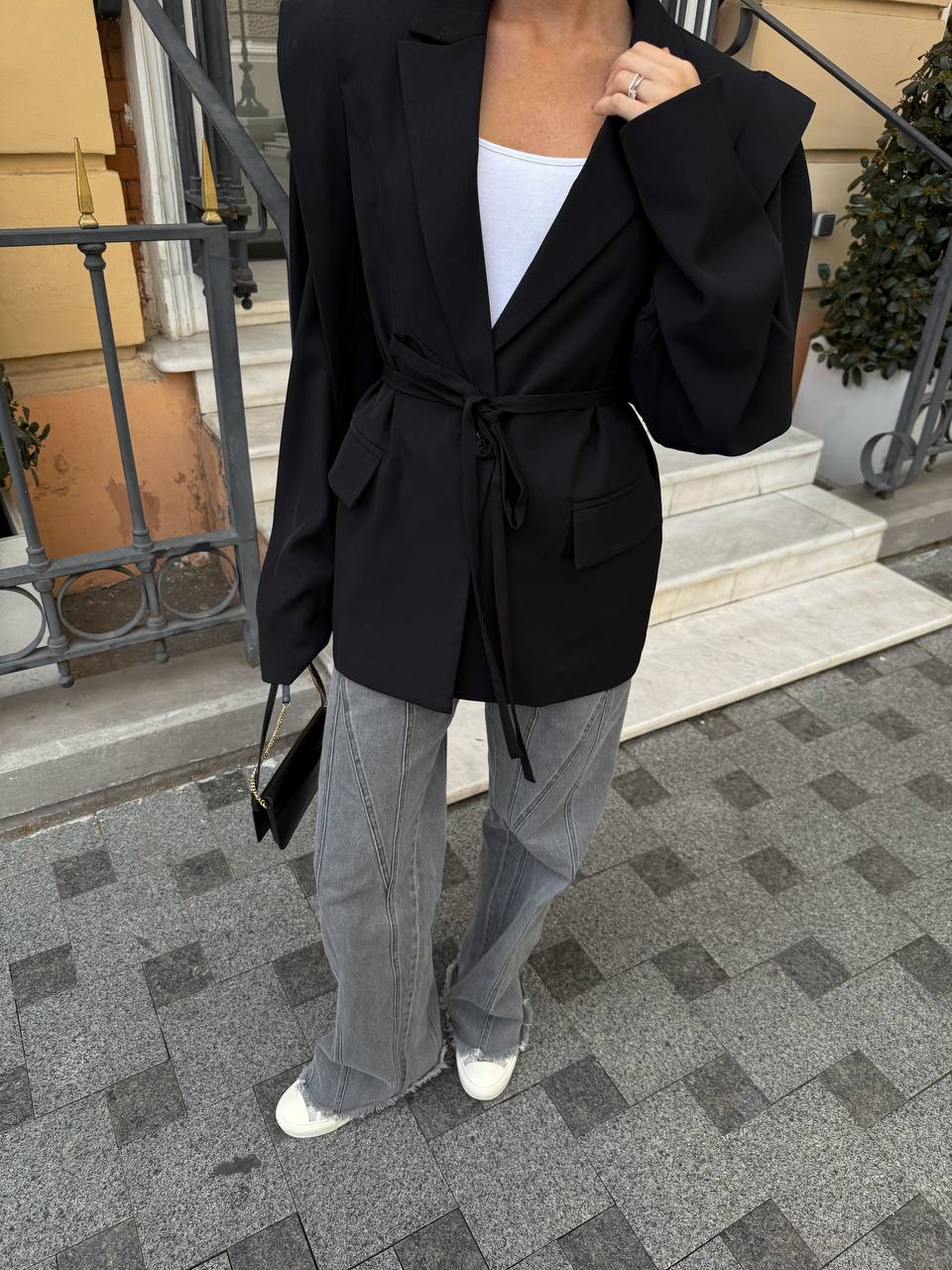 Person wearing a black coat with a tie waist, white shirt, gray pants, and white shoes on a sidewalk.