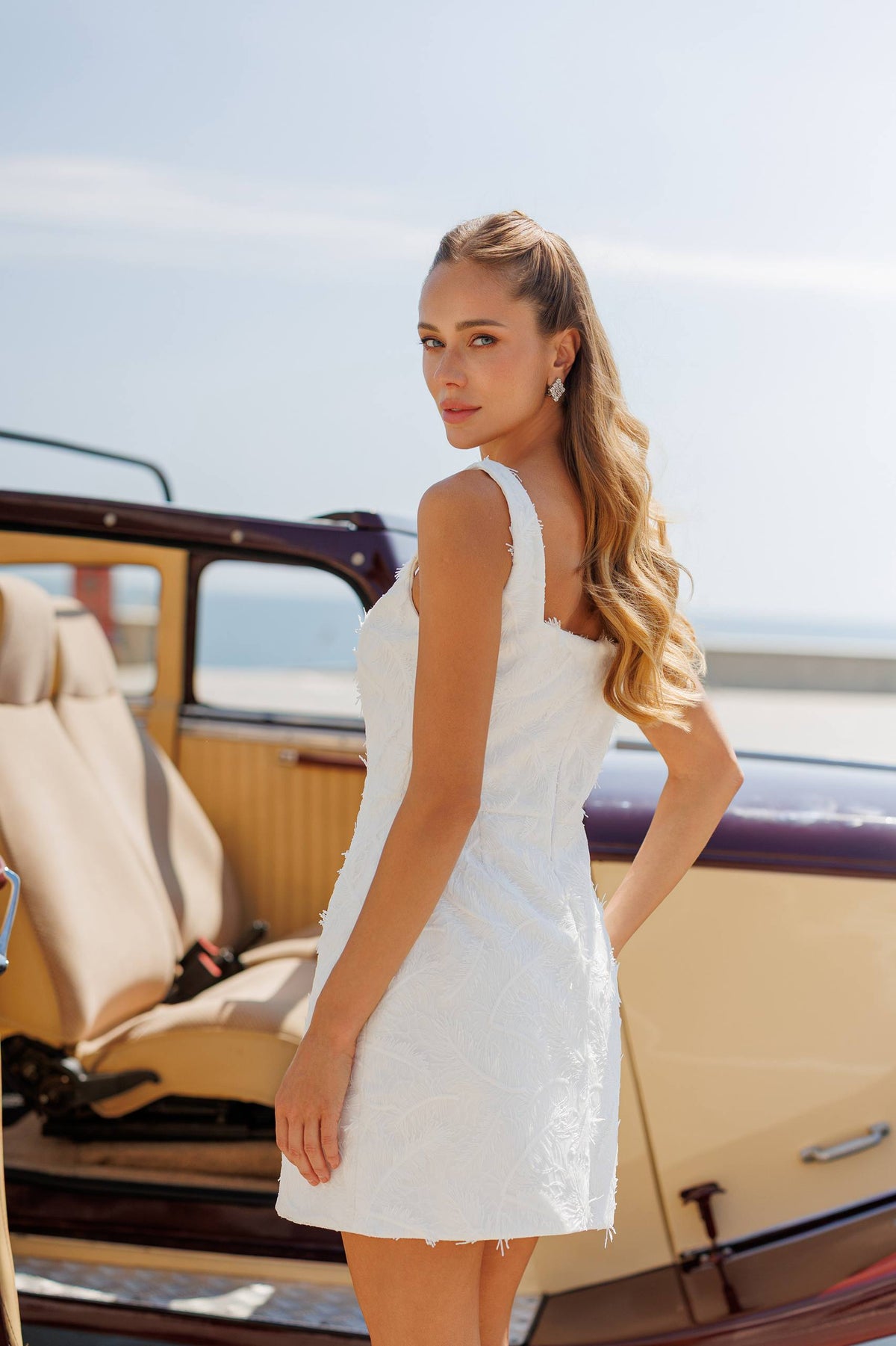 Woman in a white dress standing next to a vintage car with a clear sky background