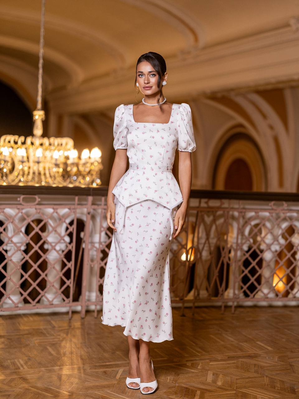 Woman in a white floral dress standing in an elegant indoor setting with chandeliers.