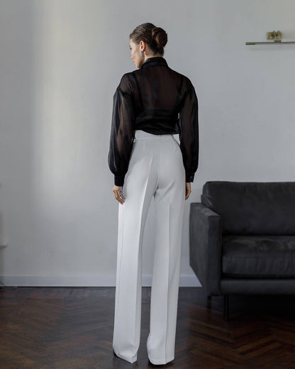 Person wearing a black sheer blouse and white pants standing in a room with a gray sofa.