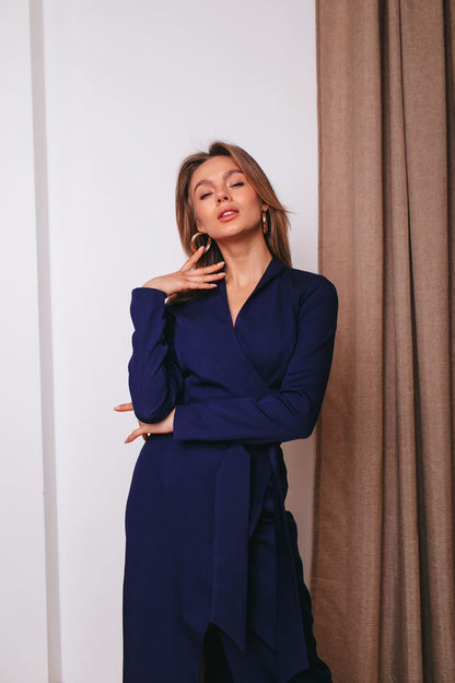 Woman wearing a blue dress standing against a white wall with a beige curtain.