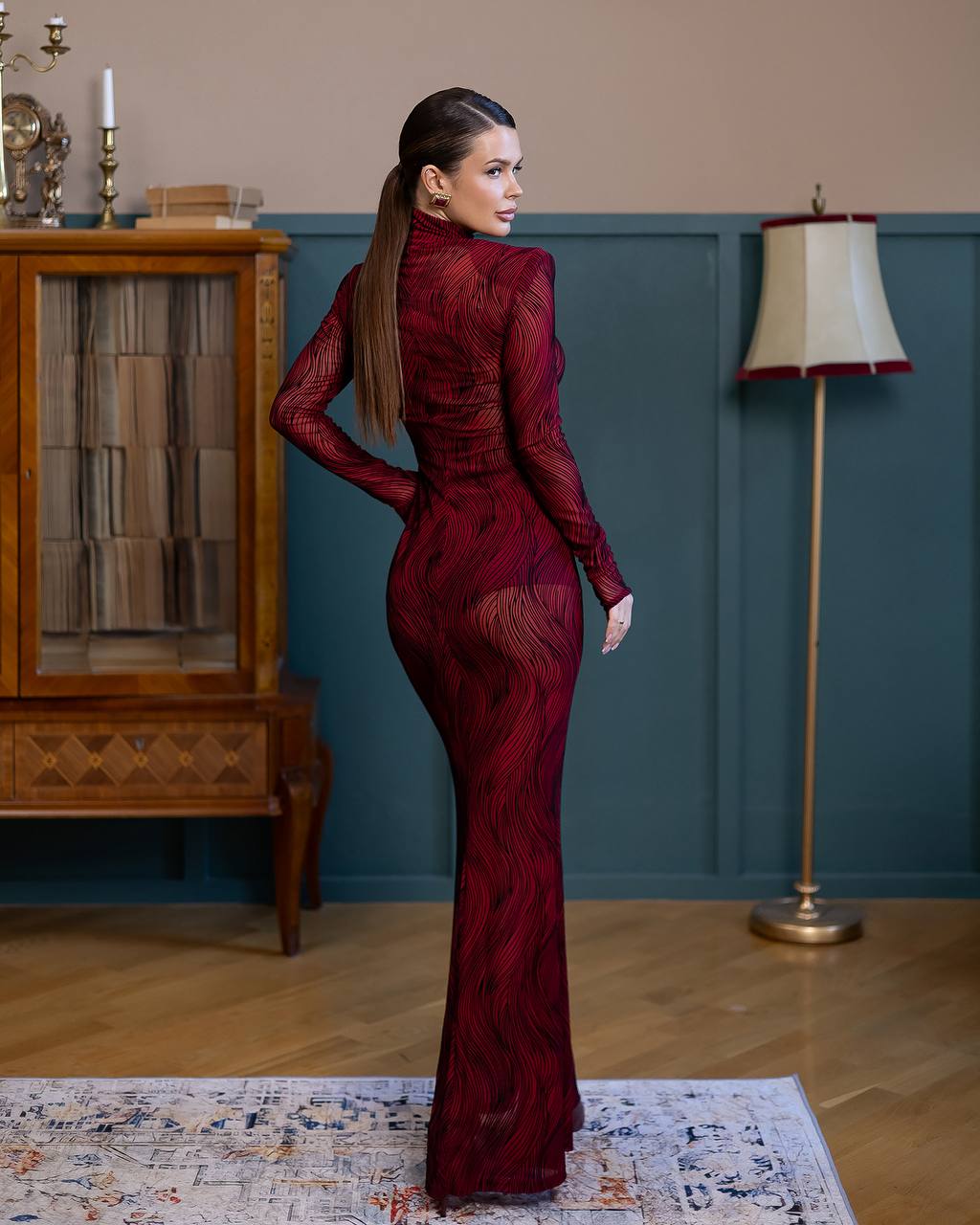 Woman in a burgundy evening gown standing in a room with wooden floor and decorative elements.