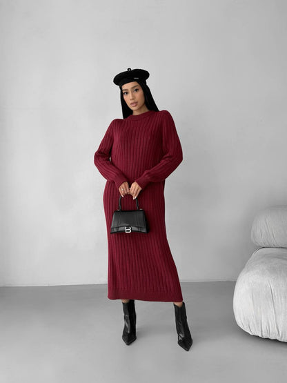 Woman wearing a burgundy dress and black boots, holding a black handbag against a plain background.