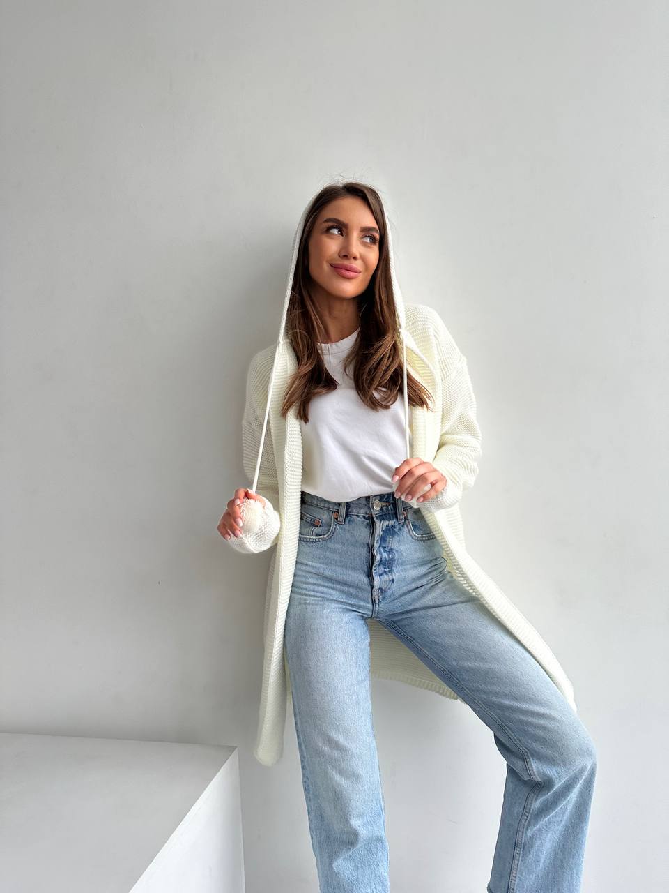 Woman wearing a long white cardigan, white shirt, and blue jeans against a plain background