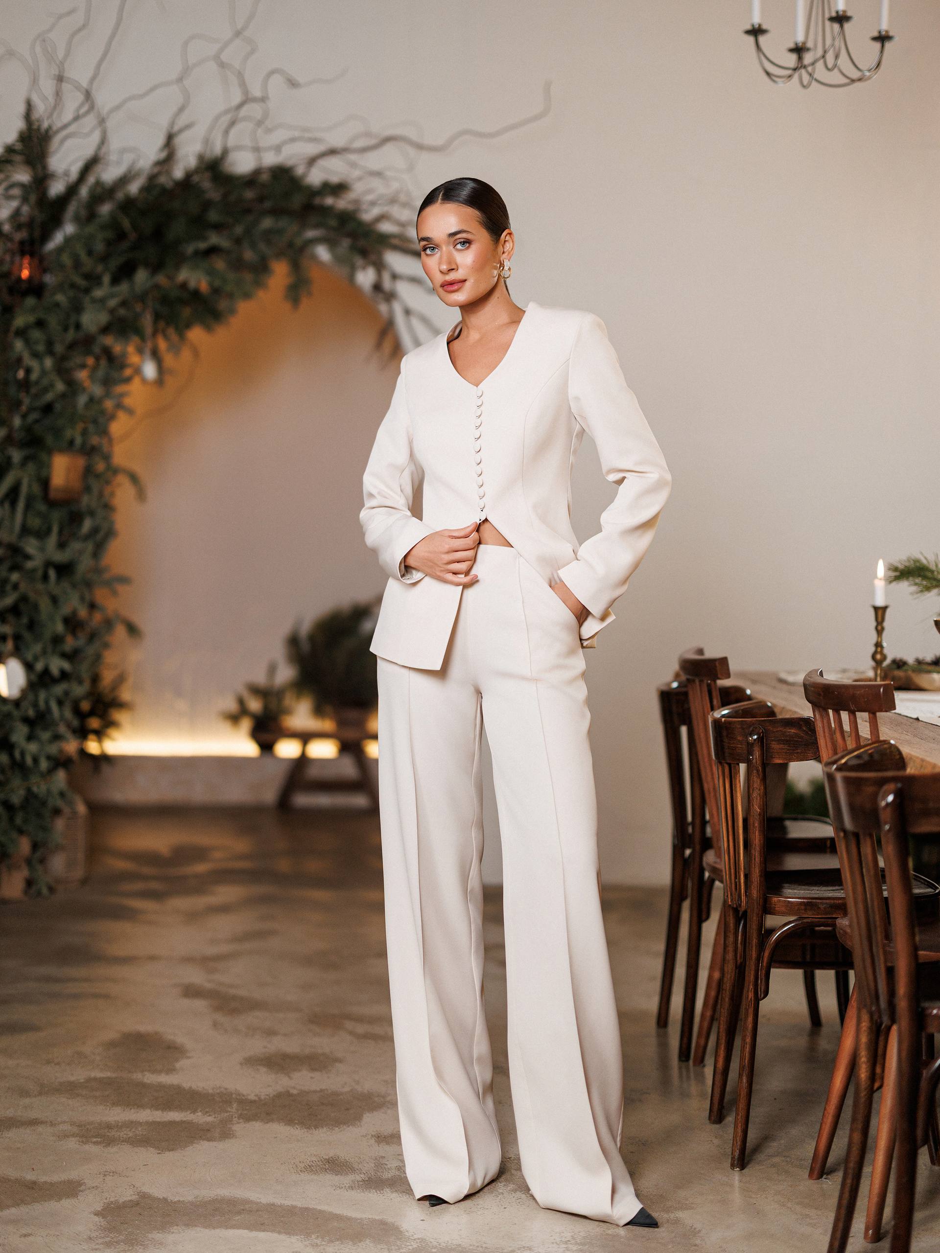 Woman in a white suit standing in a decorated room with plants and wooden furniture.