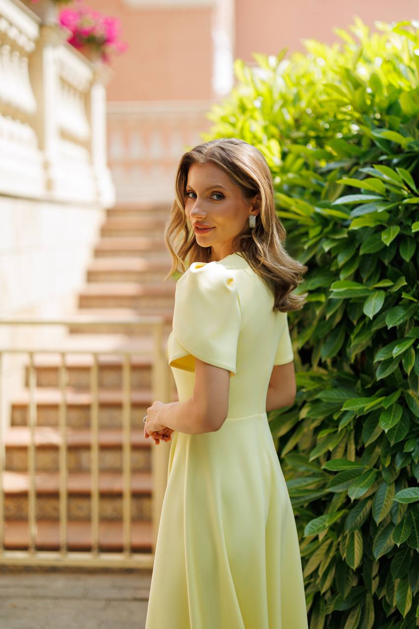 A woman wearing a pale yellow dress stands outdoors beside a green hedge and stone stairs, looking back over her shoulder.
