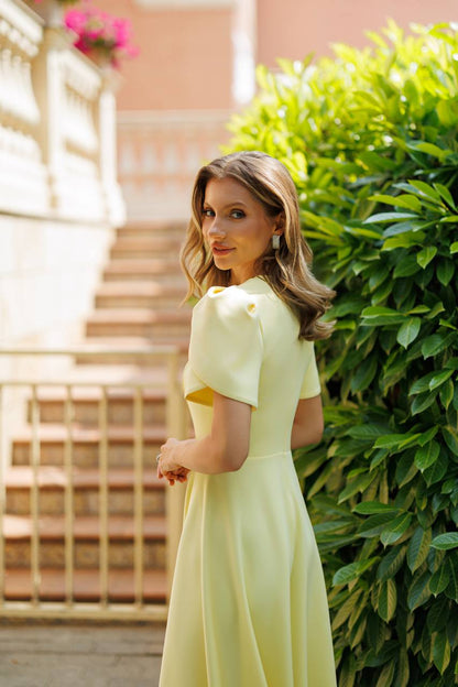 A woman wearing a pale yellow dress stands outdoors beside a green hedge and stone stairs, looking back over her shoulder.
