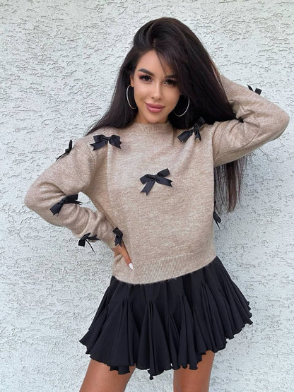 Woman wearing a beige sweater with black bows and a black skirt against a light gray wall.