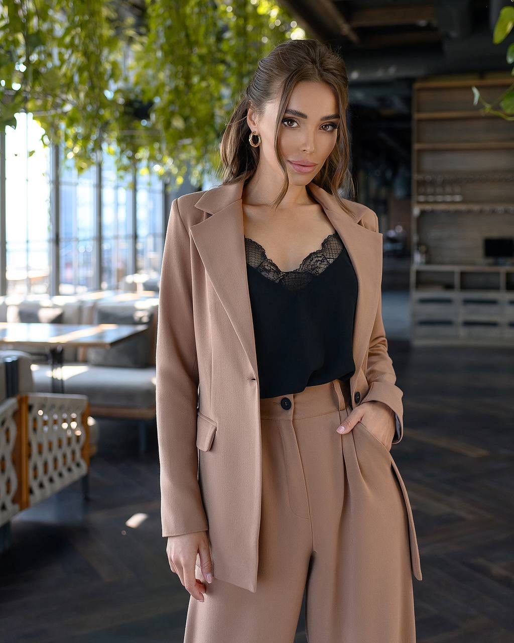 Woman wearing a beige coat over a black top in an indoor setting with plants and furniture.