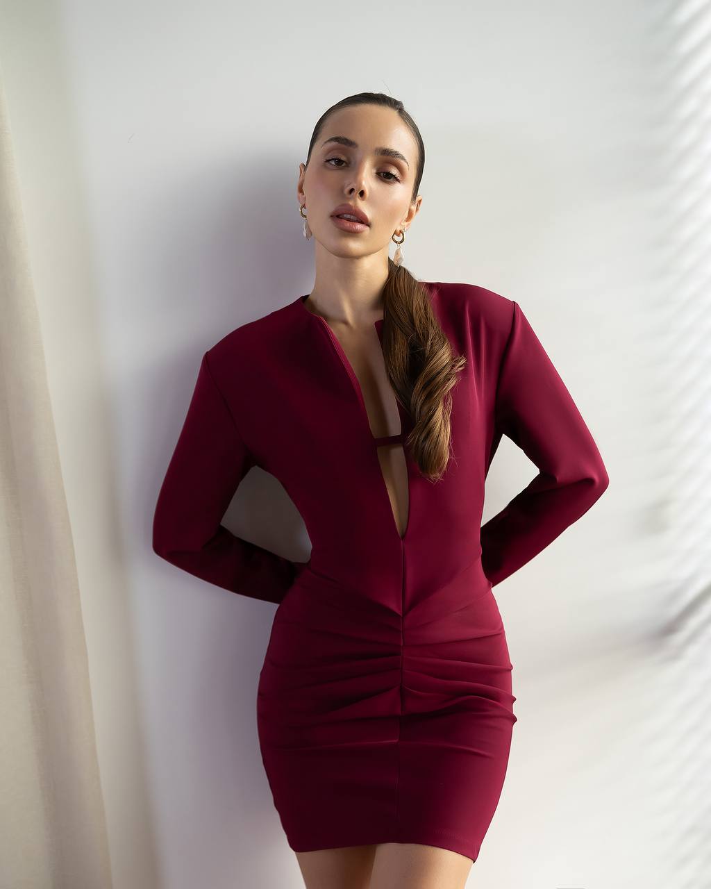 A woman wearing a fitted long-sleeve mini dress in deep burgundy with a plunging V-neckline and ruched detailing, posing against a white wall with soft window shadow patterns.