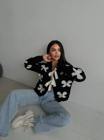 Woman wearing a black cardigan with white floral patterns and blue jeans sitting on a gray floor.