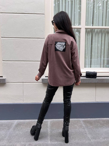 Person wearing a brown jacket with a logo on the back, standing in front of a building.