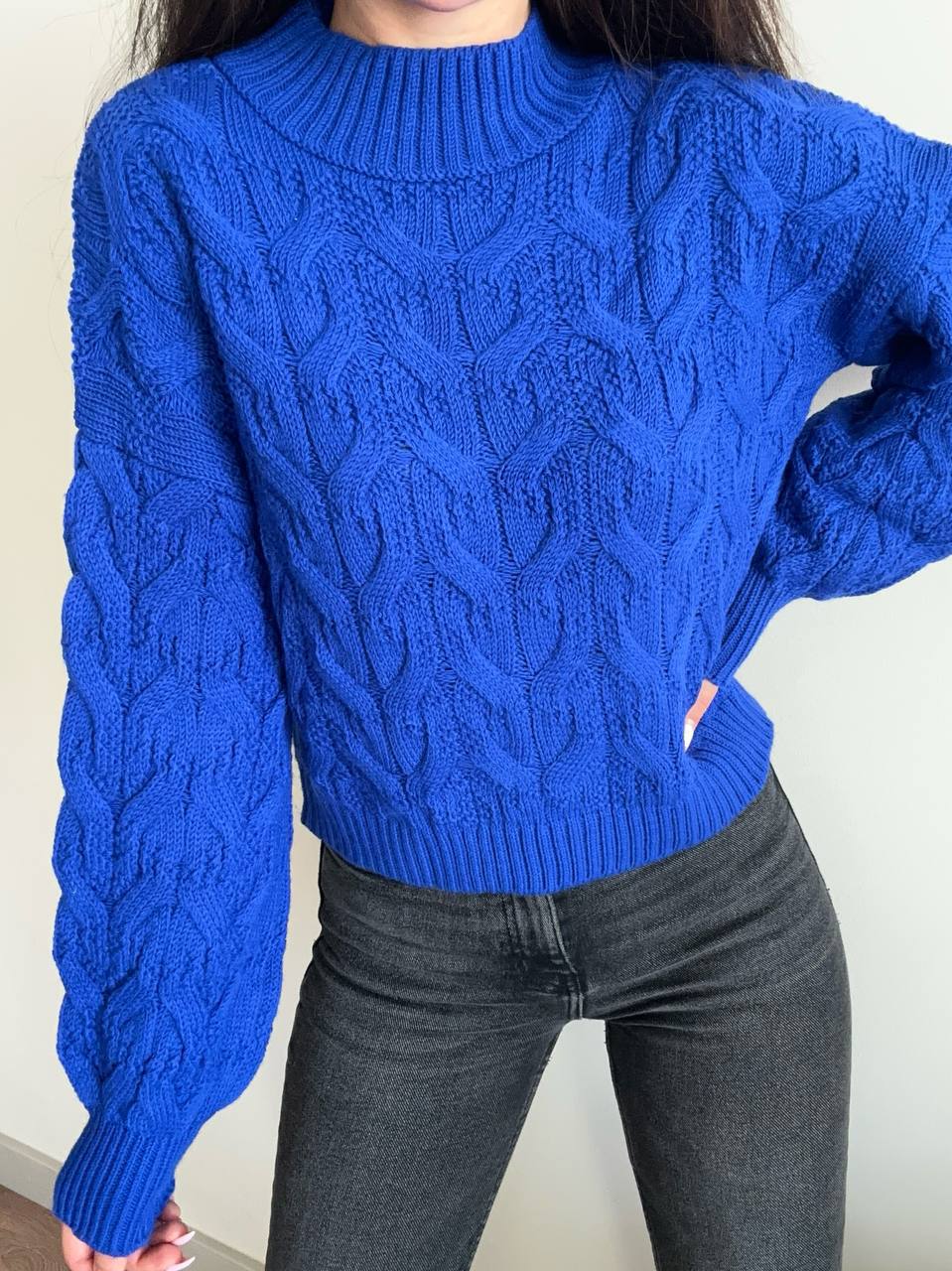 Person wearing a blue cable knit sweater and dark jeans against a neutral background